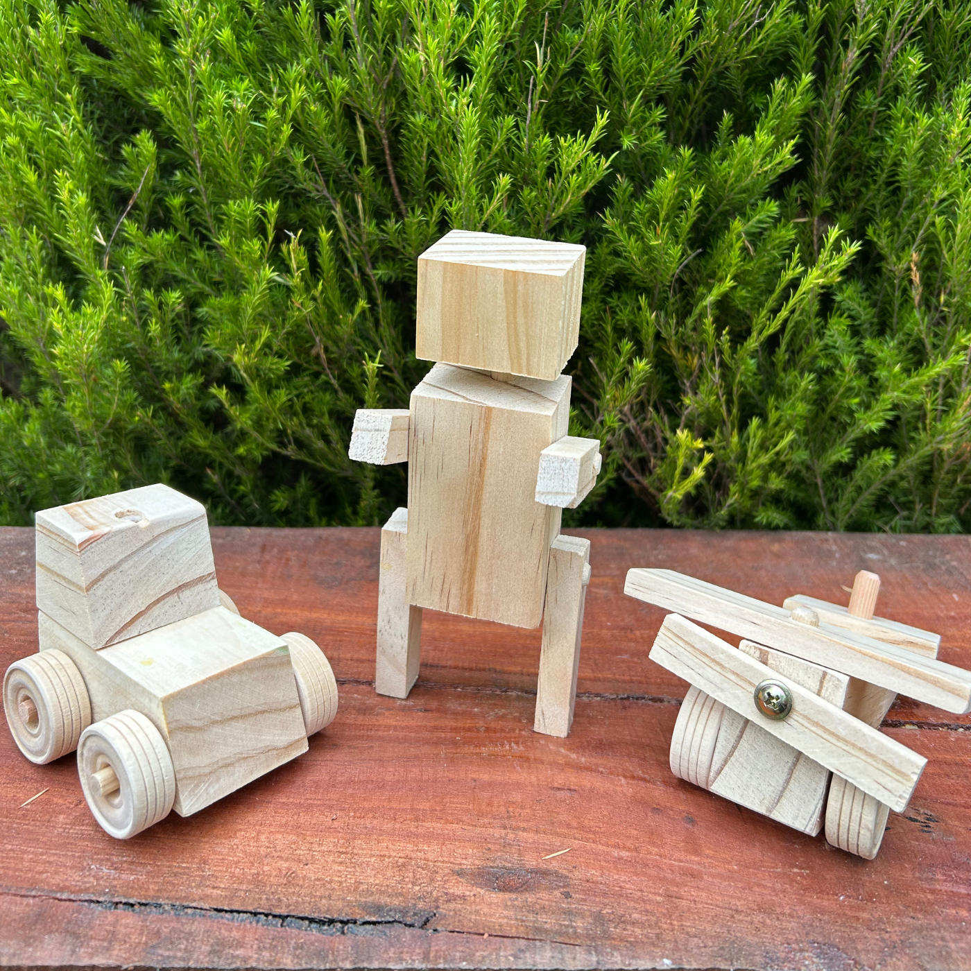Three wooden toys including a car, robot, and airplane on a wooden surface with greenery in the background.