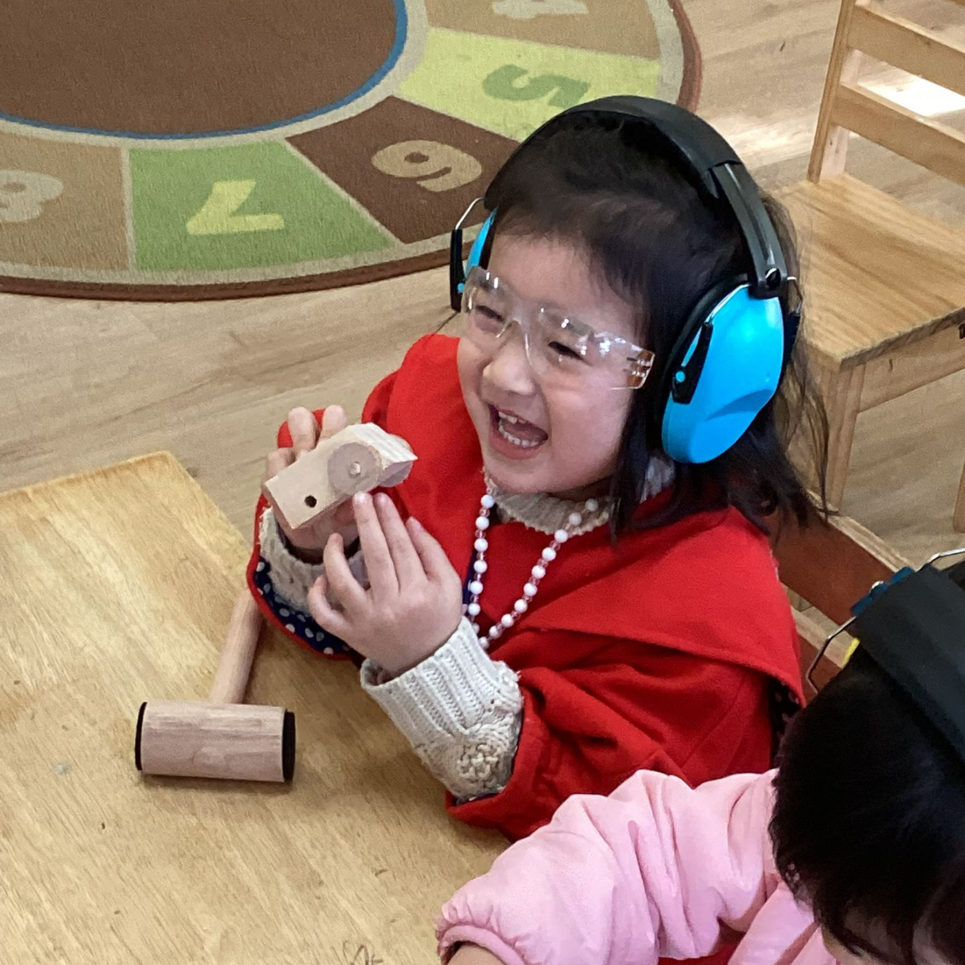 Child wearing headphones and safety goggles, holding a wooden tool on a wooden floor.
