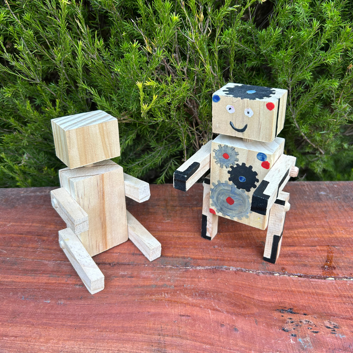 Two wooden robot toys on a wooden surface with greenery in the background